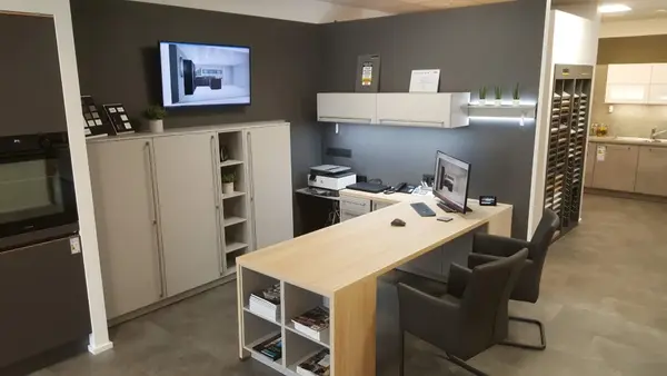 Modernes Küchenstudio-Büro mit Schreibtisch, Computer und Musterküchen im Hintergrund.